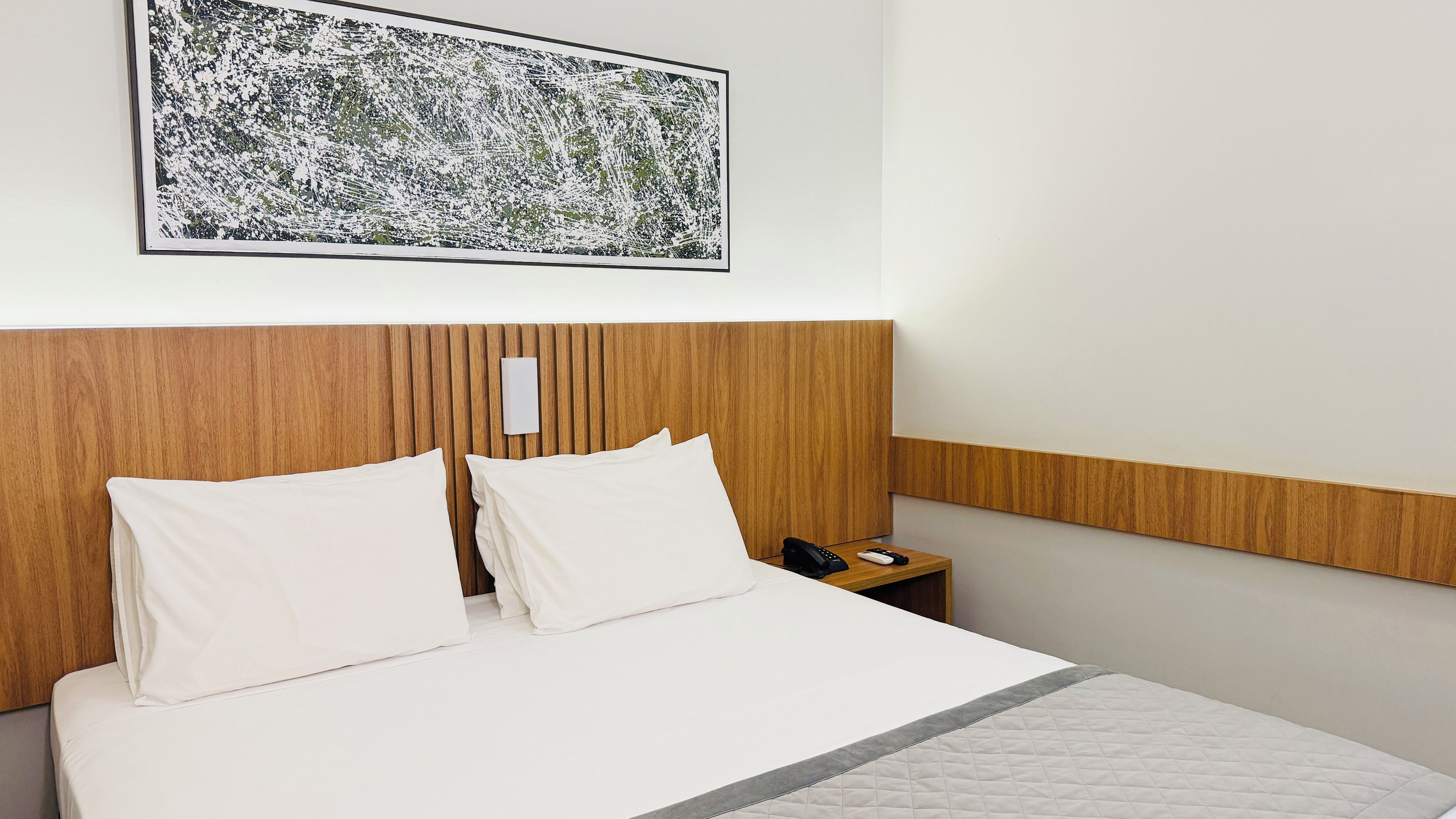 Habitación de hotel con cabecera de madera, cama blanca con dos almohadas, mesita de noche con teléfono y una obra de arte abstracta enmarcada en la pared.