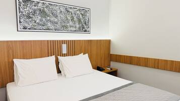 Habitación de hotel con cabecera de madera, cama blanca con dos almohadas, mesita de noche con teléfono y una obra de arte abstracta enmarcada en la pared.