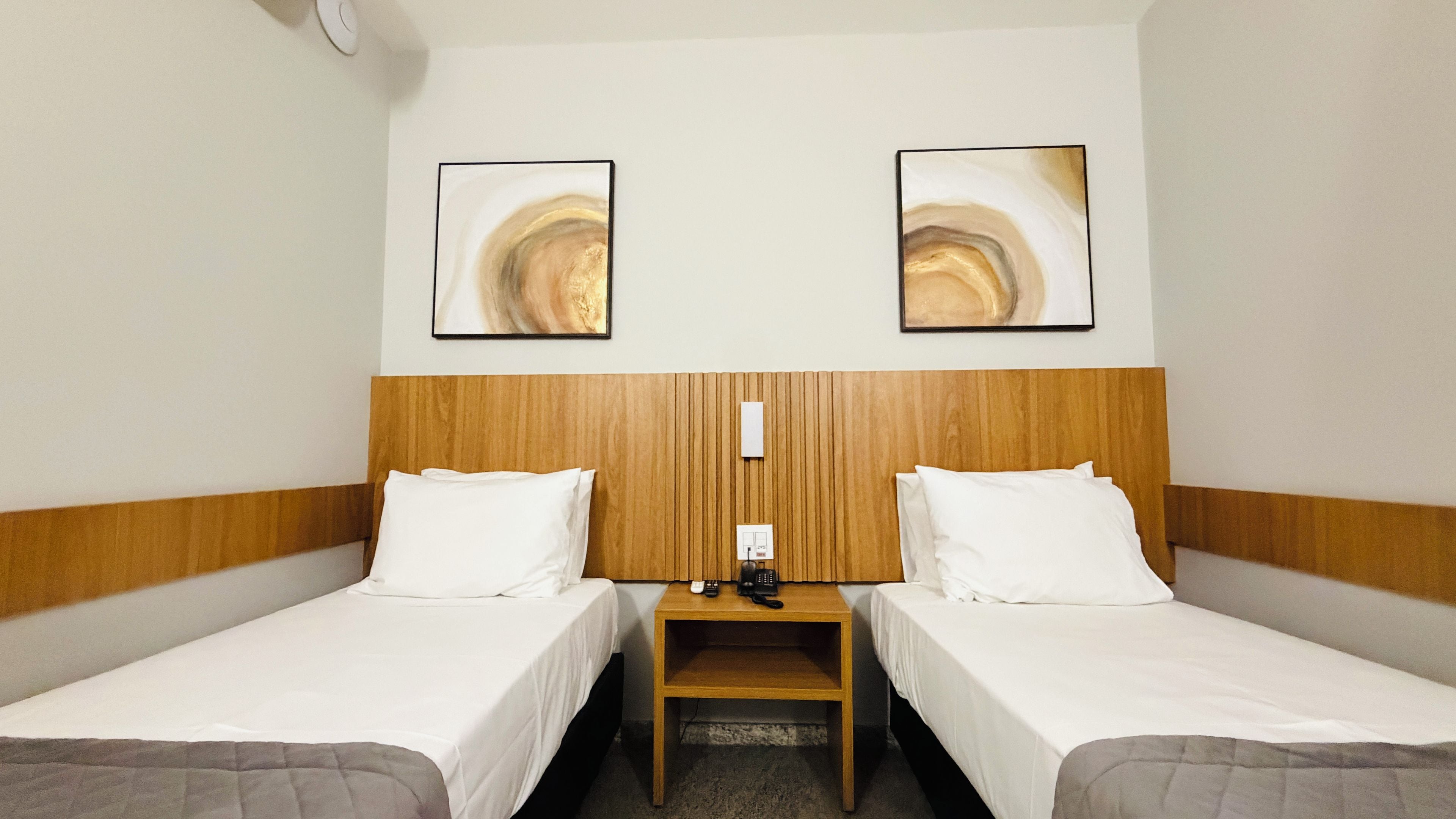 A hotel room with two single beds, a wooden headboard, a nightstand with a phone, and abstract artwork on the walls.