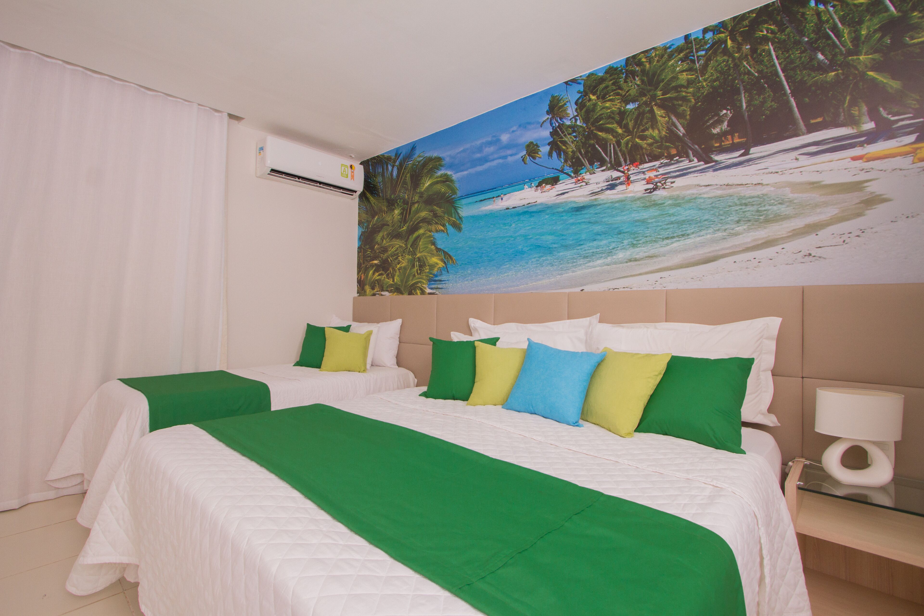 A hotel room with two beds, a beach mural, a white air conditioner, and a bedside table with a lamp.