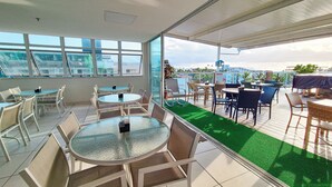 A rooftop terrace with round tables and chairs, large windows, and a view of the cityscape. - Pousada Marie Claire Flats (Ipojuca)