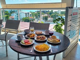 Un tavolo rotondo con prodotti per la colazione, tra cui pane tostato, frutta, uova strapazzate e succo, posizionato su un balcone con vista su edifici e palme.