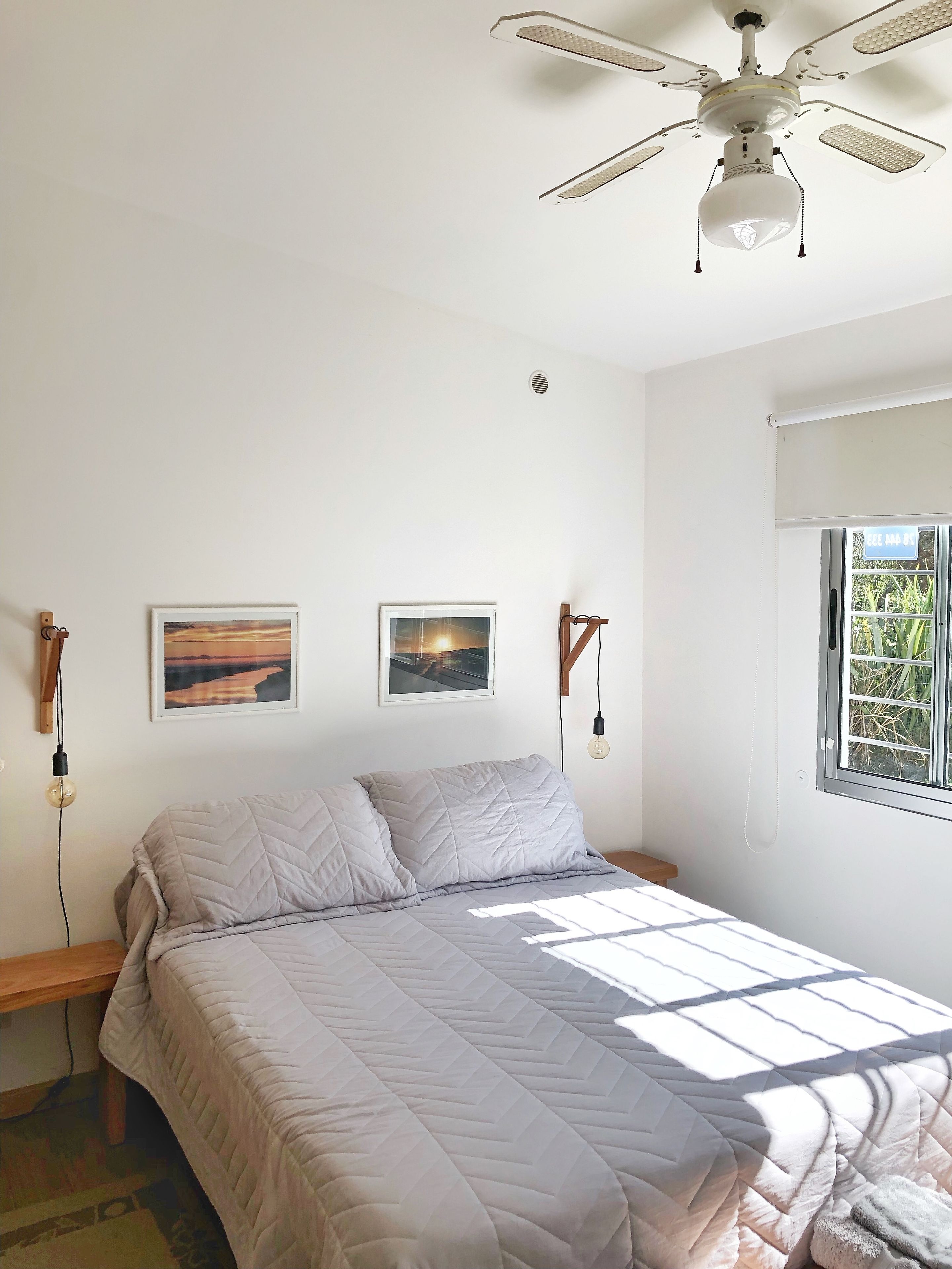 A bedroom with a bed, ceiling fan, two wall-mounted light fixtures, and a window with a view of greenery.