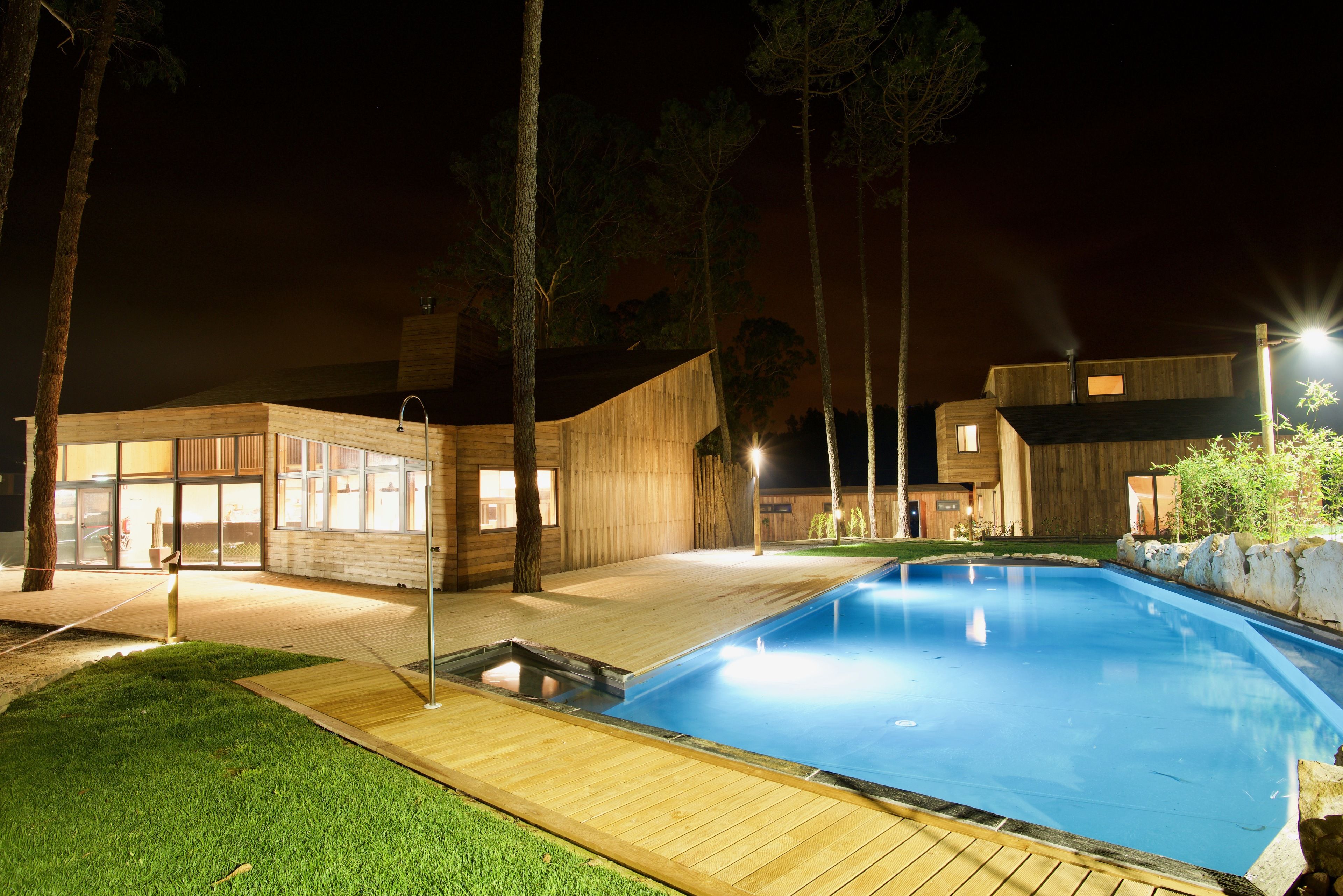A modern outdoor pool area with a wooden deck, a small hot tub, and a well-lit building with large windows.