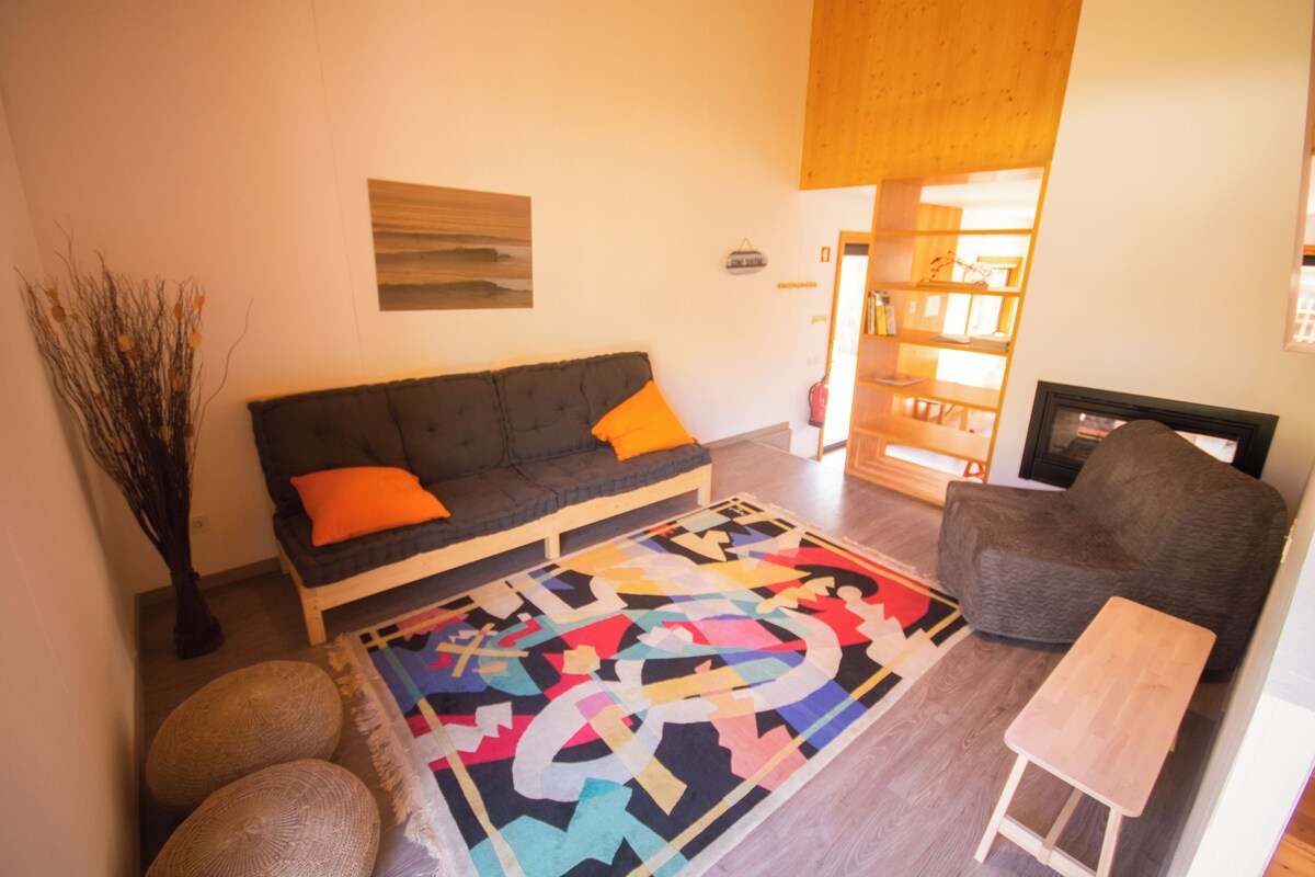 A living room with a dark grey sofa, orange cushions, a colorful abstract rug, and a small wooden table.