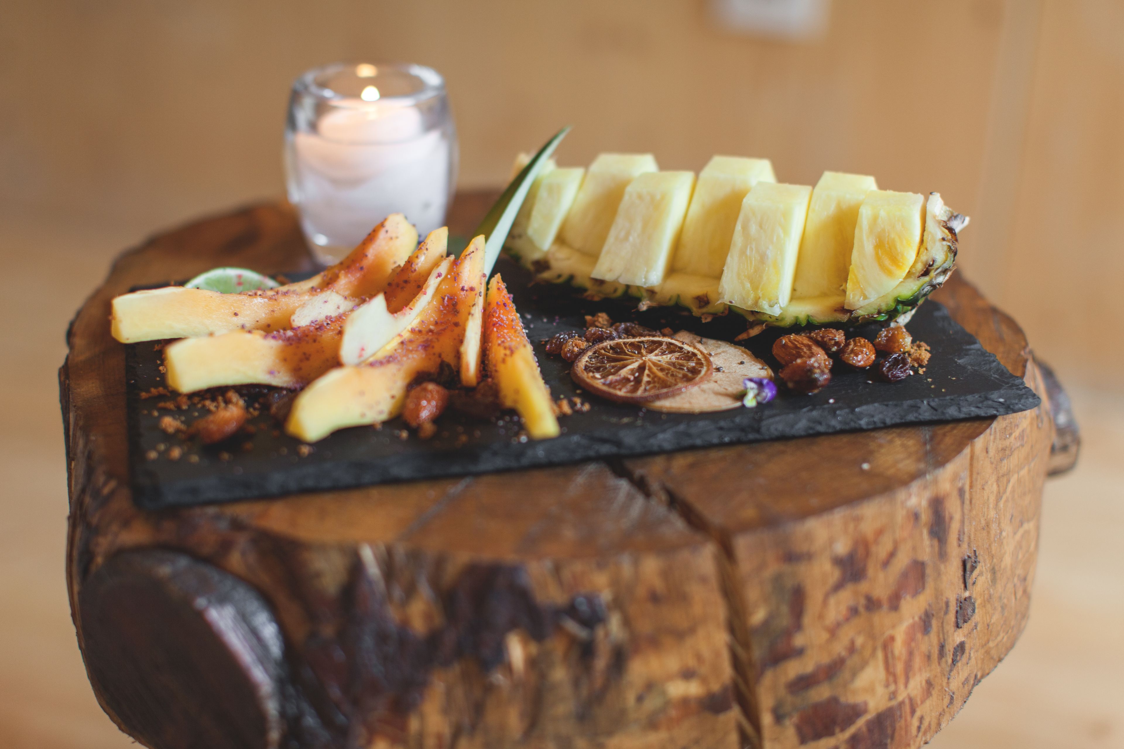 Une table en bois sur laquelle repose un plateau noir en ardoise, servant de présentoir pour des fruits coupés, des noix et une bougie.