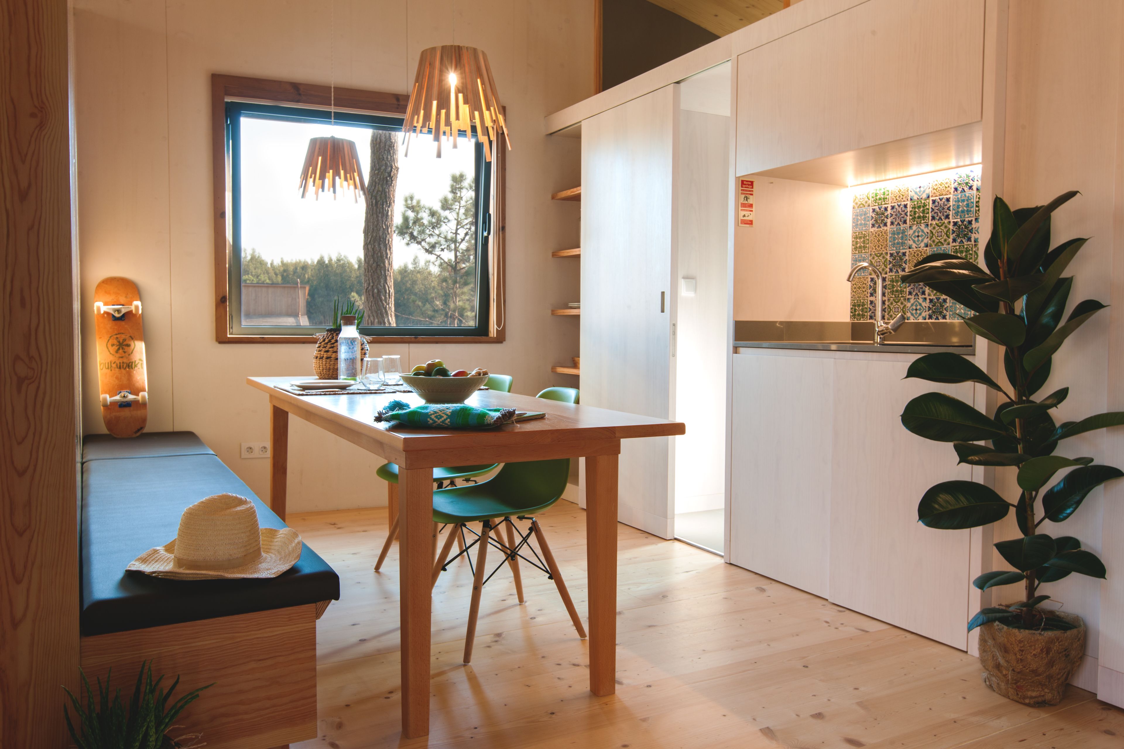 A modern kitchen with a wooden table, chairs, a hanging light fixture, and a view of the outdoors through a window.