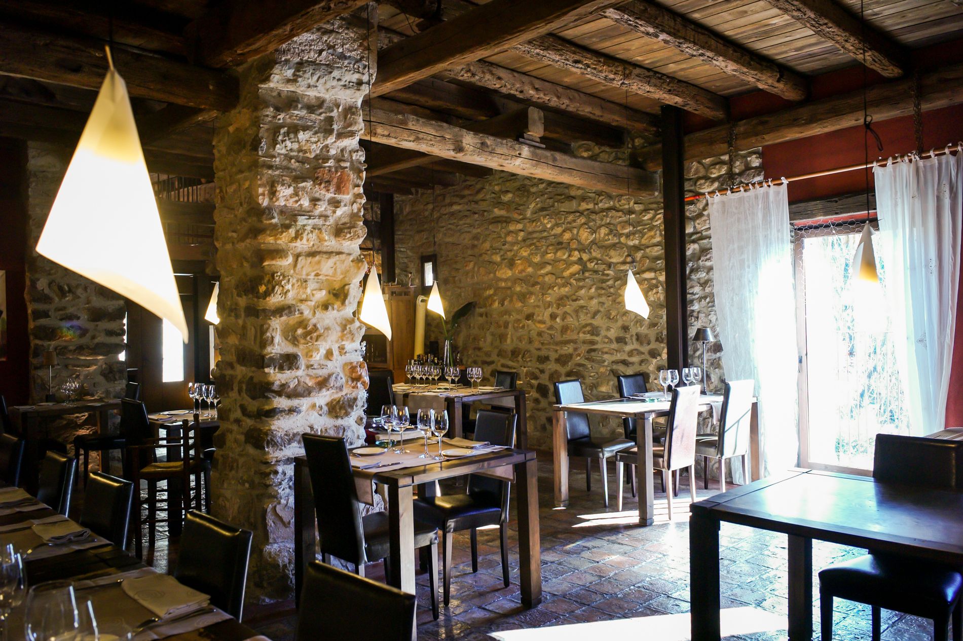 A rustic dining area with stone walls, wooden beams, and hanging pendant lights. Tables are set with wine glasses and plates.