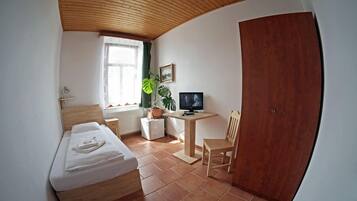 Une petite pièce bien éclairée, avec un lit, un bureau équipé d’un ordinateur, une chaise, une plante en pot et une armoire en bois.