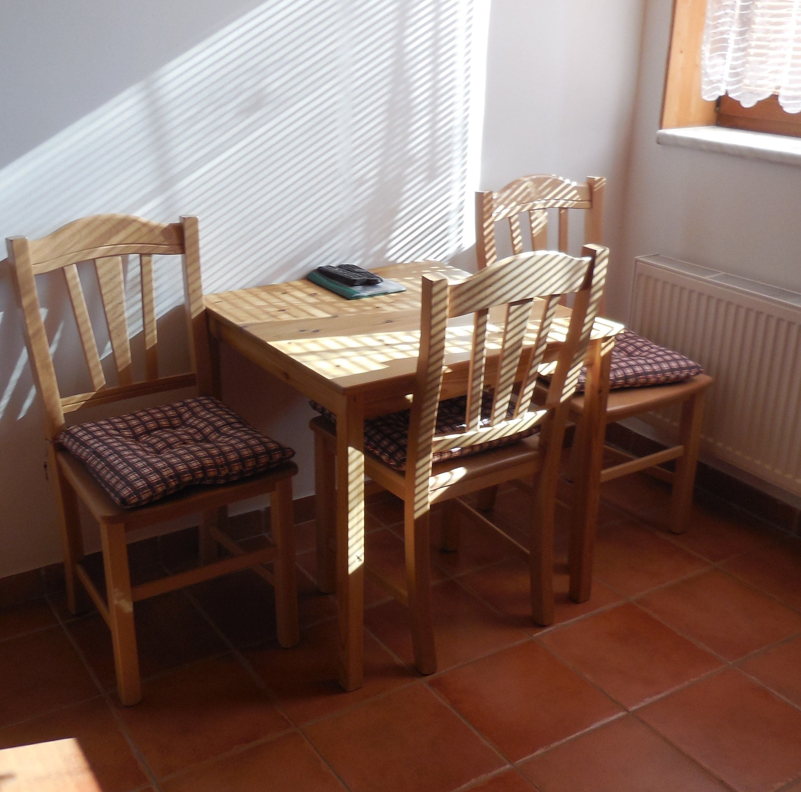 Une table à manger en bois avec des chaises, une télécommande posée sur la table et, à l’arrière-plan, une fenêtre avec des stores.