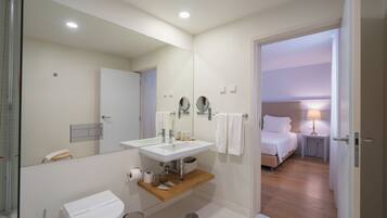 Un baño moderno con lavamanos, espejo y inodoro blancos. Hay un estante de madera debajo del lavamanos y un pequeño cubo de basura. Una puerta da acceso a un dormitorio con cama y mesita de noche con lámpara.