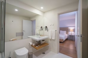 Un bagno moderno con lavandino, specchio e servizi igienici bianchi. Sotto il lavandino si trova una mensola in legno e un cestino. Una porta conduce a una camera da letto con un letto e un comodino con una lampada.
