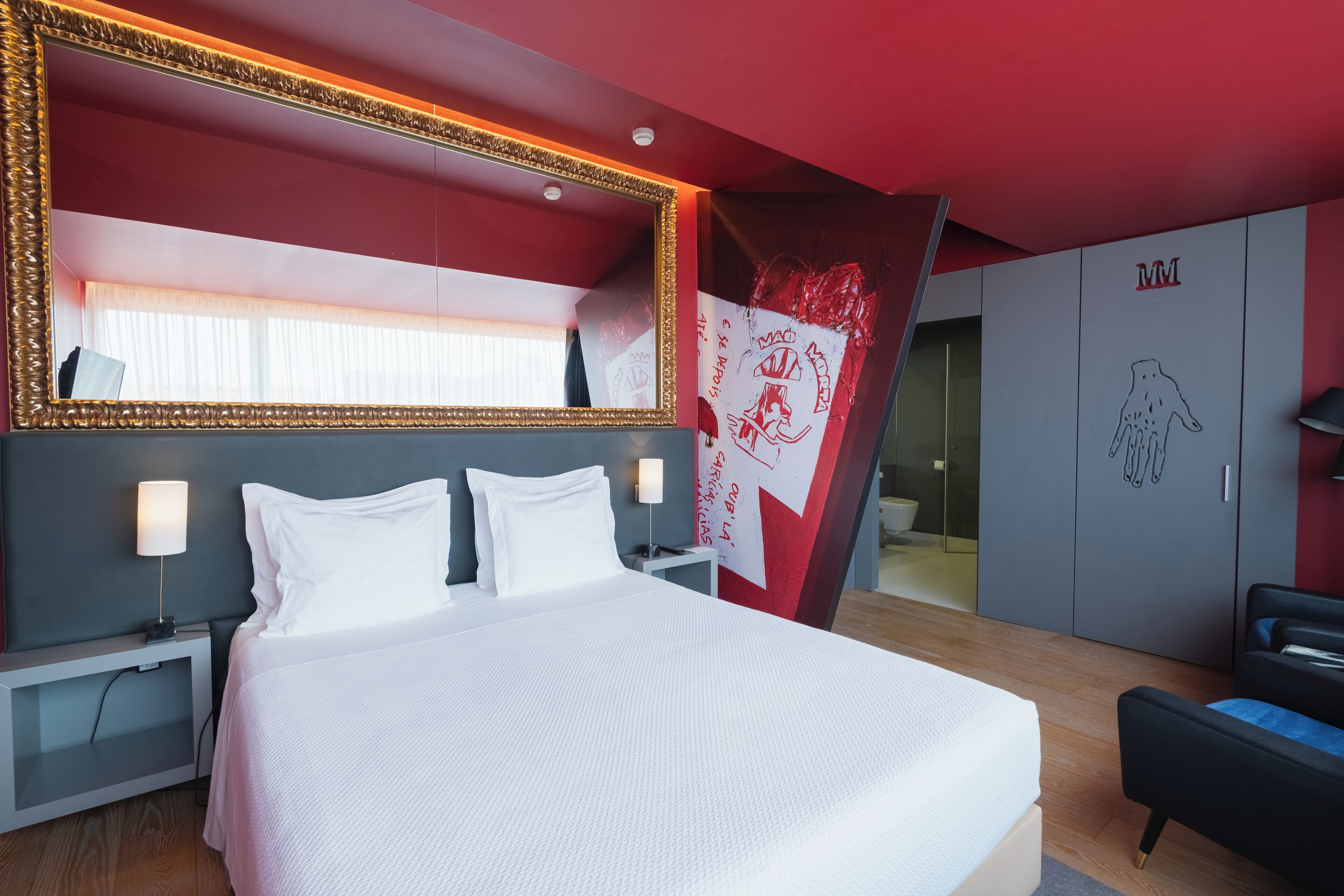 A modern hotel room with a large bed, a bedside table with a lamp, a wardrobe, and a large mirror with a decorative frame.