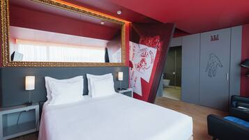 A modern hotel room with a large bed, a bedside table with a lamp, a wardrobe, and a large mirror with a decorative frame.