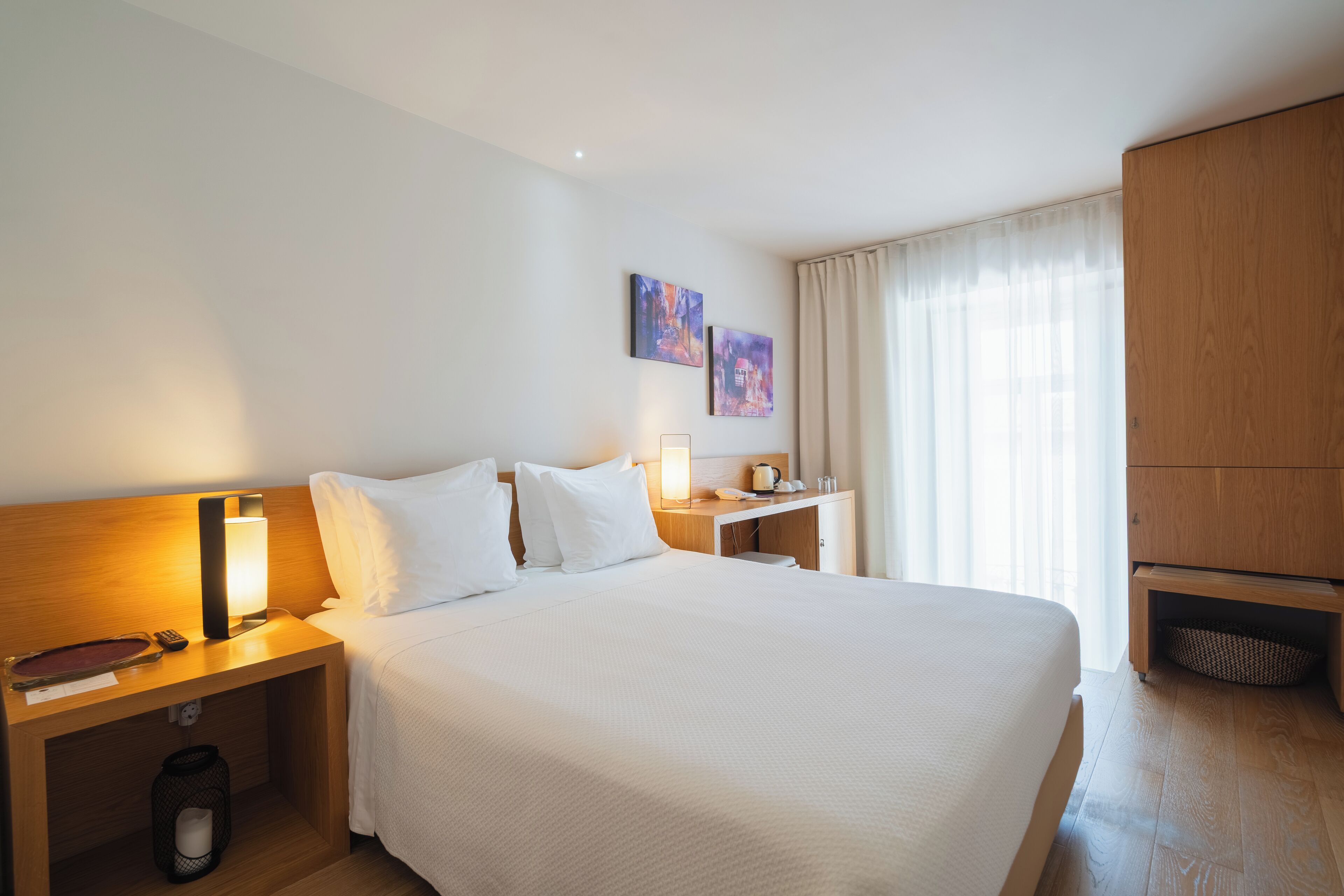A hotel room with a large bed, a nightstand with a lamp, a wooden wardrobe, and two paintings on the wall.