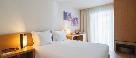 A hotel room with a large bed, a nightstand with a lamp, a wooden wardrobe, and two paintings on the wall.