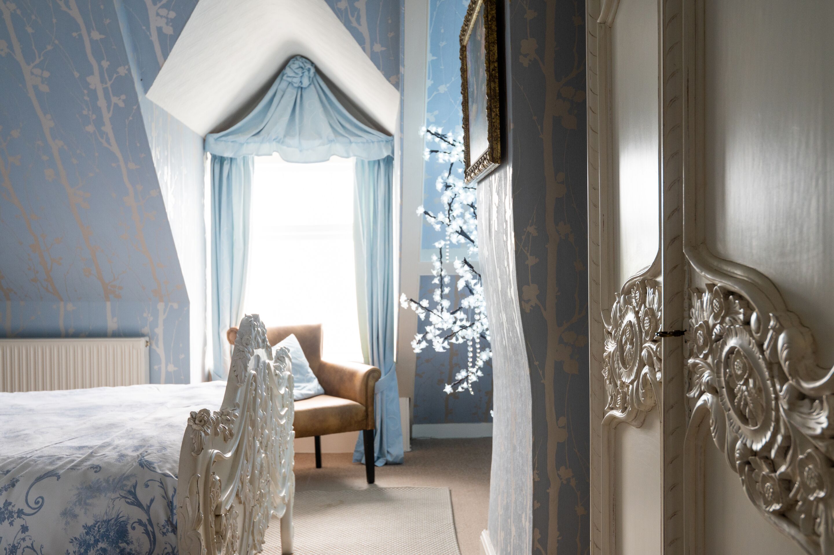 Een slaapkamer met een groot bed, een stoel, een raam met gordijnen en decoratieve wandversieringen.