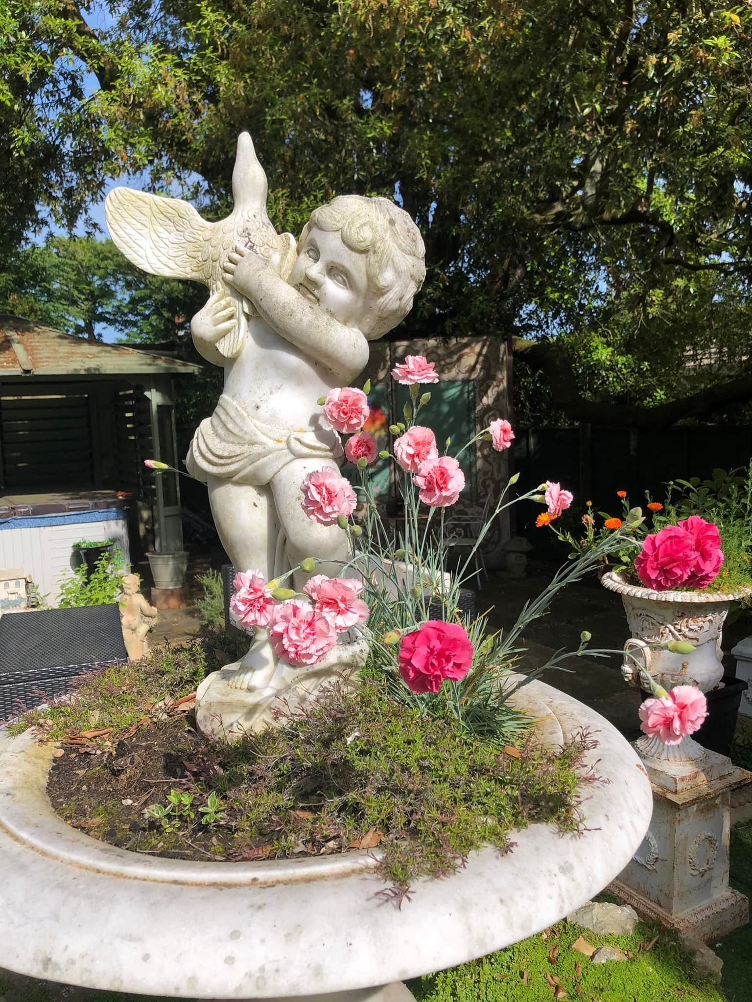 Una estatua de jardín de un ángel con alas, rodeada de flores rosadas y follaje verde.