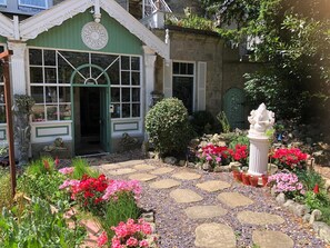 Un bâtiment pittoresque à la façade verdoyante, doté d’un chemin en pierre et agrémenté de diverses fleurs et plantes.