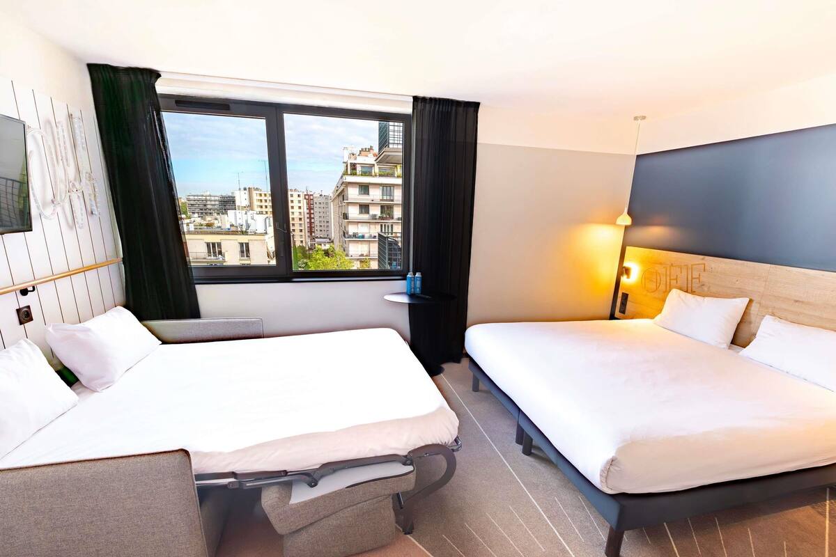 Een hotelkamer met twee bedden, een groot raam met uitzicht op de stad, een nachtkastje met een lamp en een aan de muur bevestigde televisie.