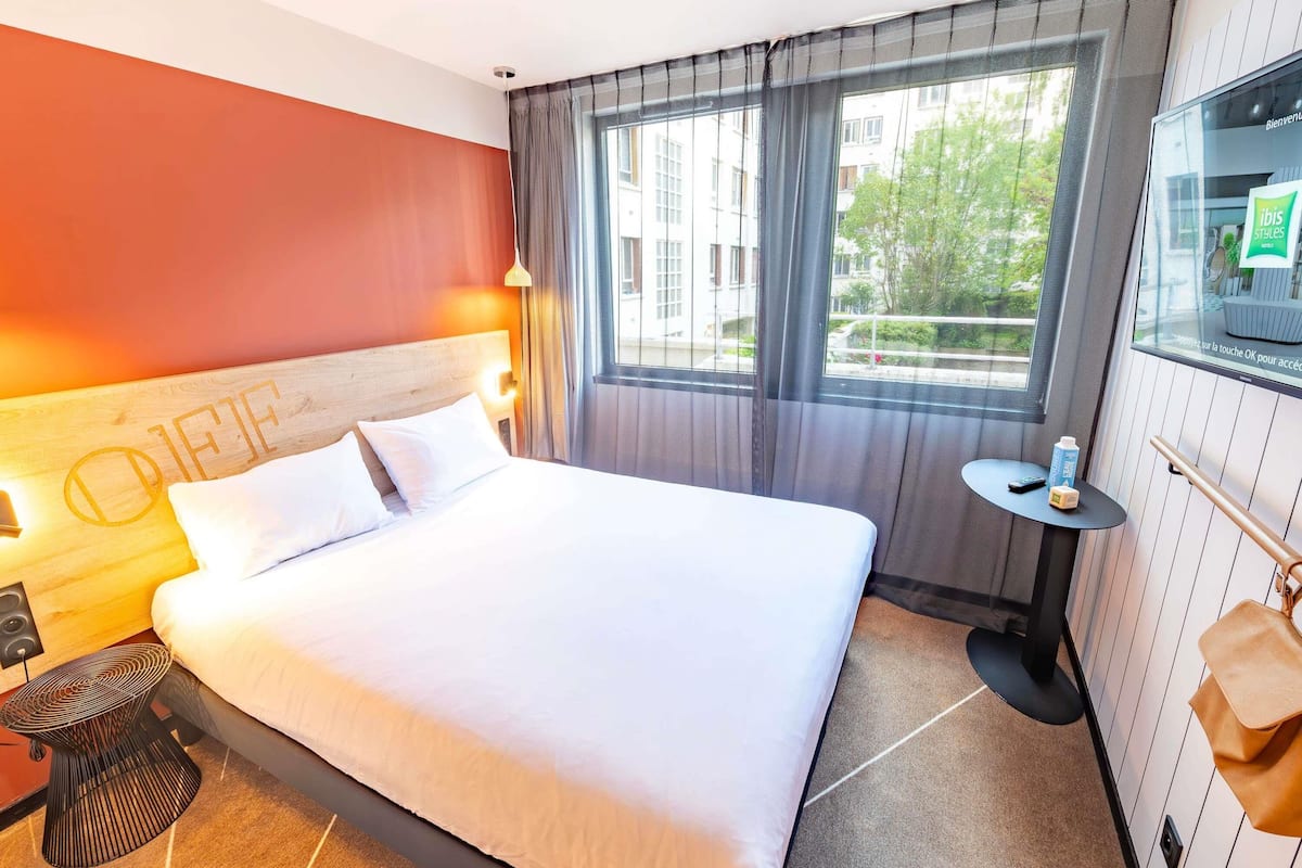 Een hotelkamer met een bed, een nachtkastje, een klein tafeltje, een raam met gordijnen en een aan de muur bevestigde televisie.