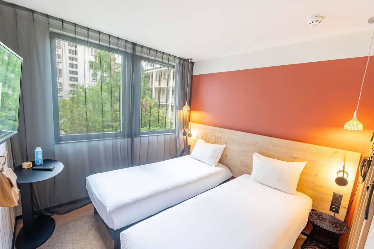 Hotelkamer met twee bedden, een kleine tafel, een stoel, een raam met gordijnen en uitzicht op gebouwen.