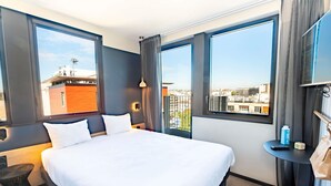 Hotelkamer met een groot bed, twee ramen, een kleine tafel, een stoel en een televisie op een standaard.