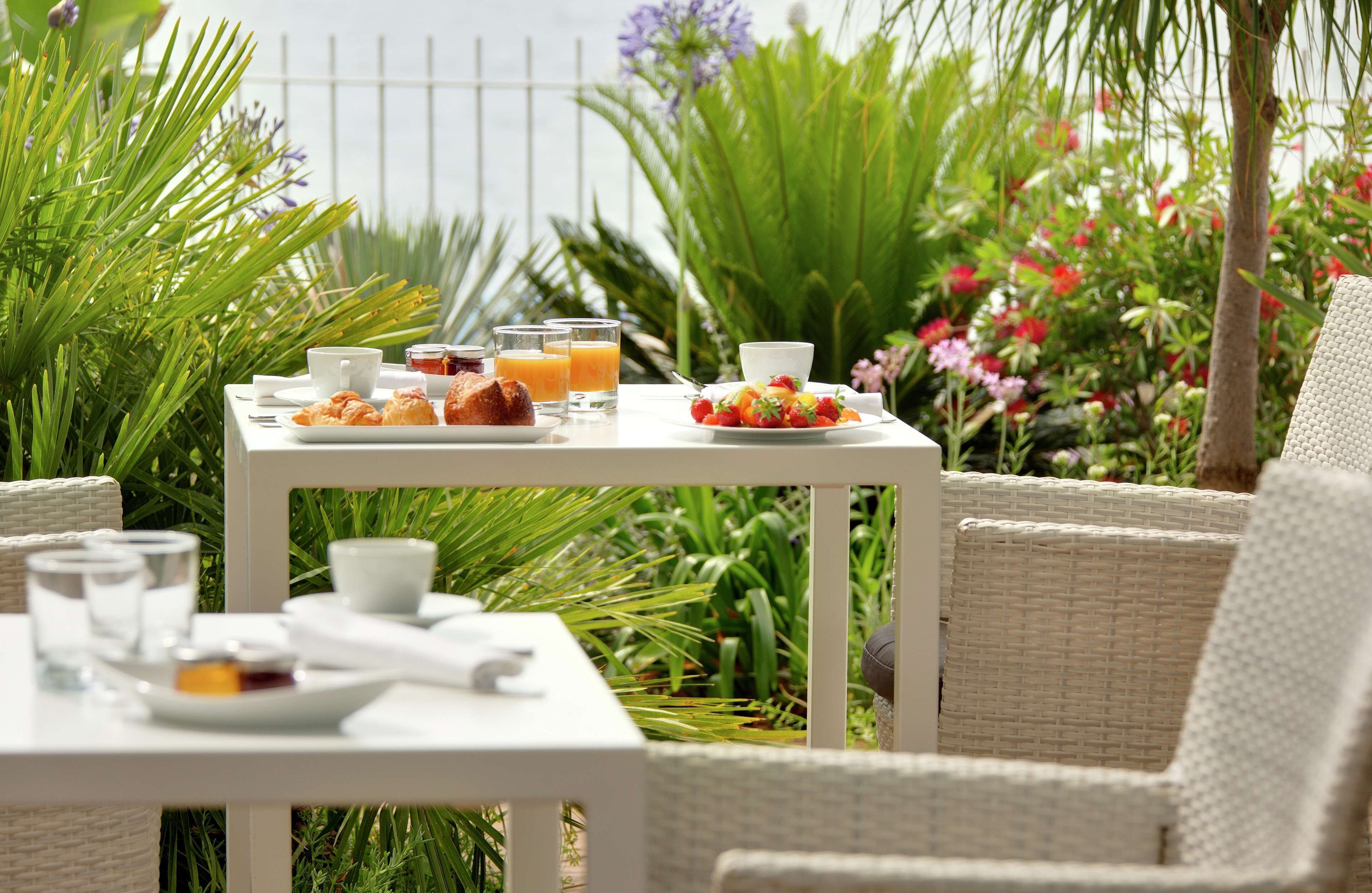 A table set with breakfast items, including croissants, fruit, and juice, surrounded by lush greenery and flowers.