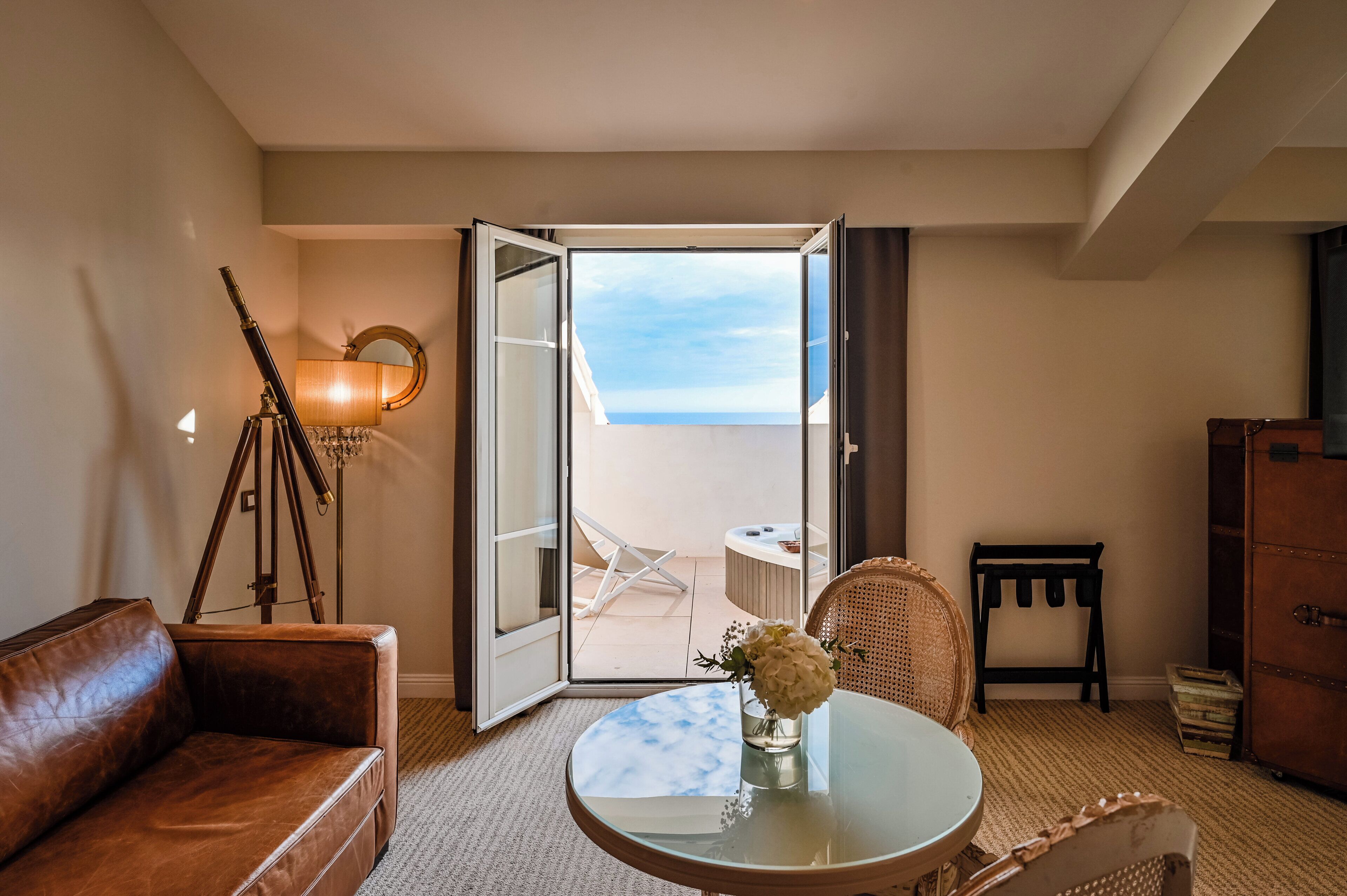 Ein Hotelzimmer mit Ledersofa, einem kleinen runden Tisch mit einer Vase, einer Dreibeinlampe und Blick aufs Meer durch eine offene Tür.