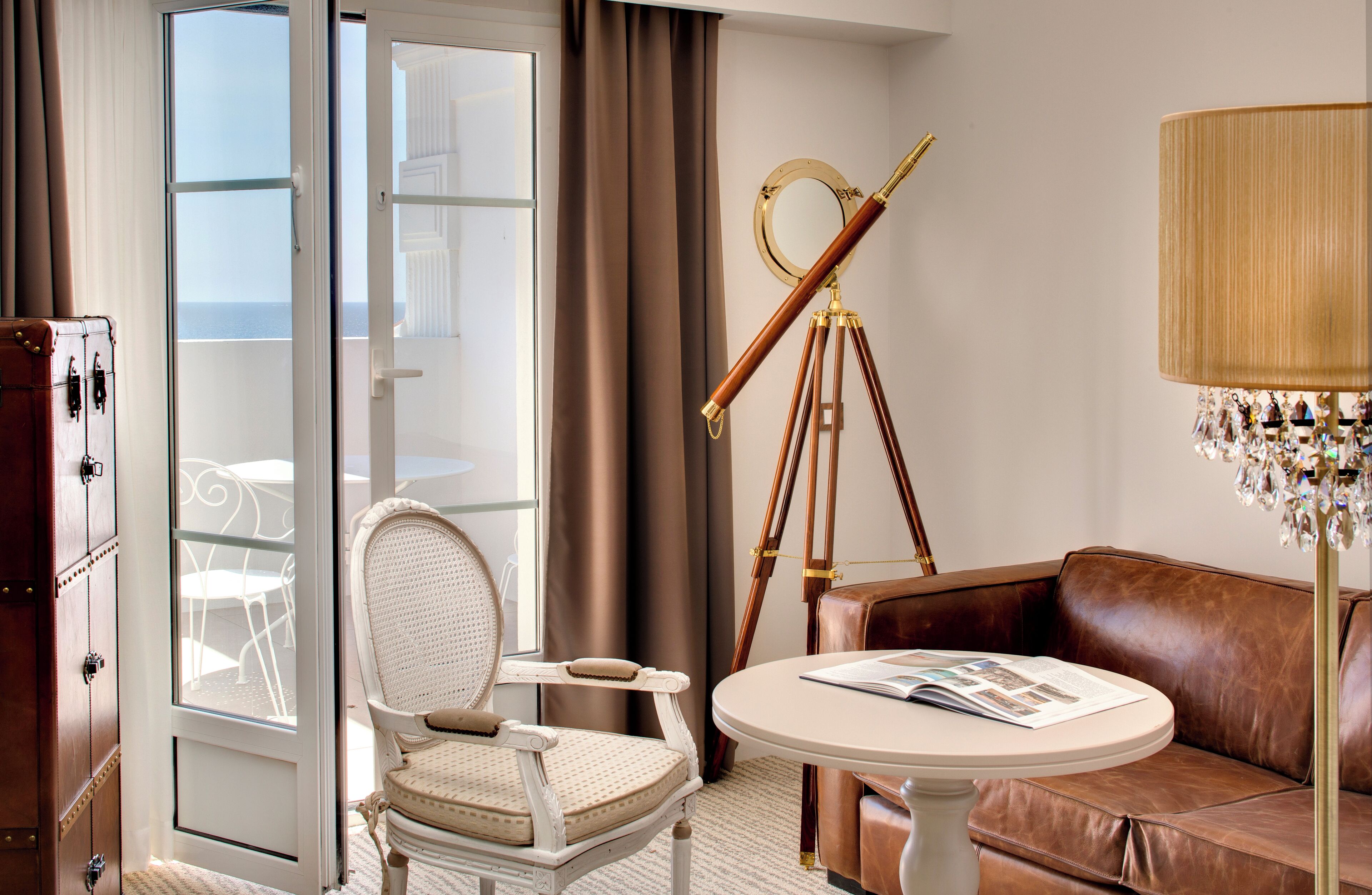 A room with a white chair, a brown leather sofa, a round table with a magazine, a telescope, and a lamp with crystals.