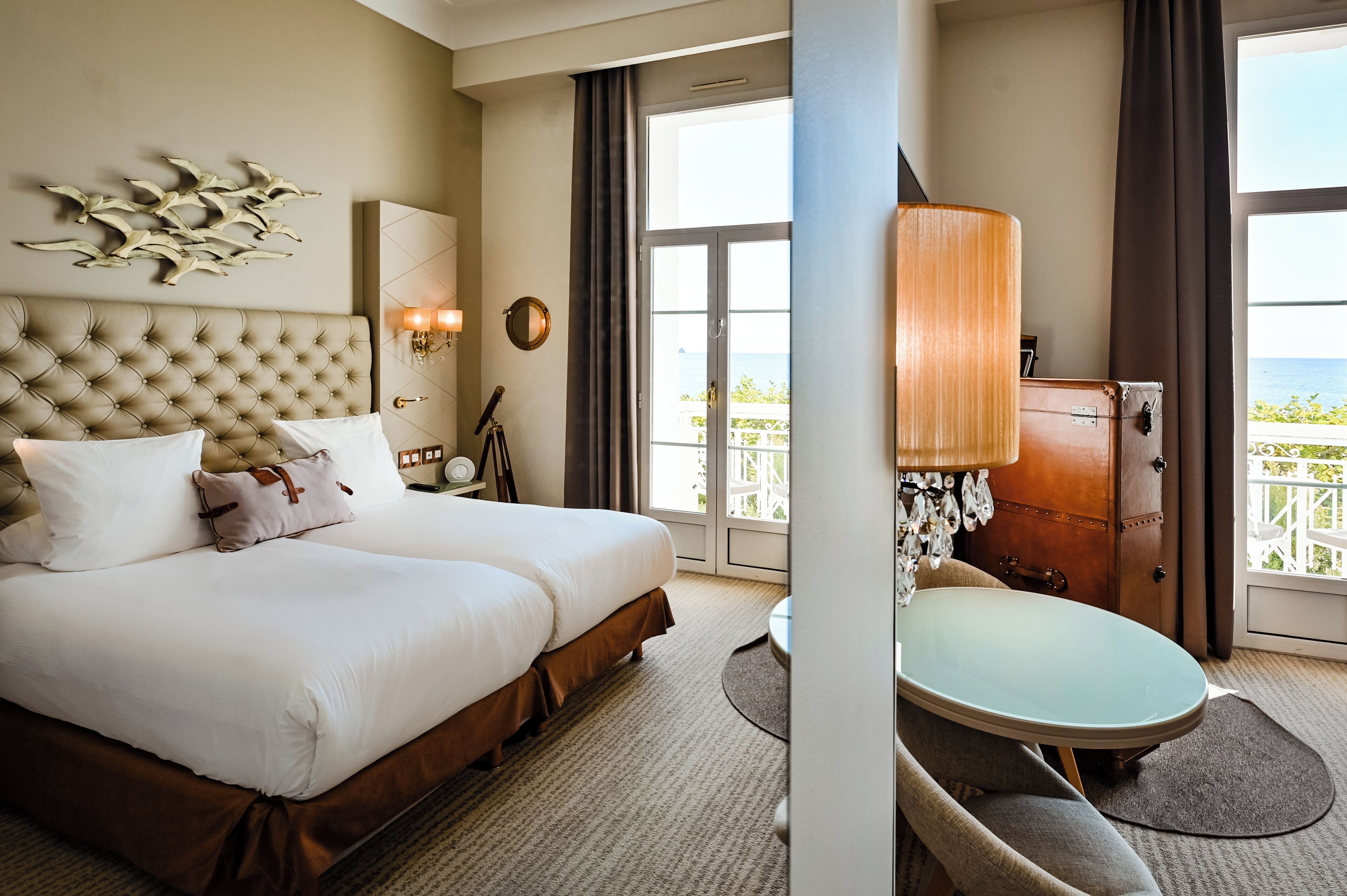 A hotel room with a large bed, a small table, a lamp, a wardrobe, and a view of the sea.
