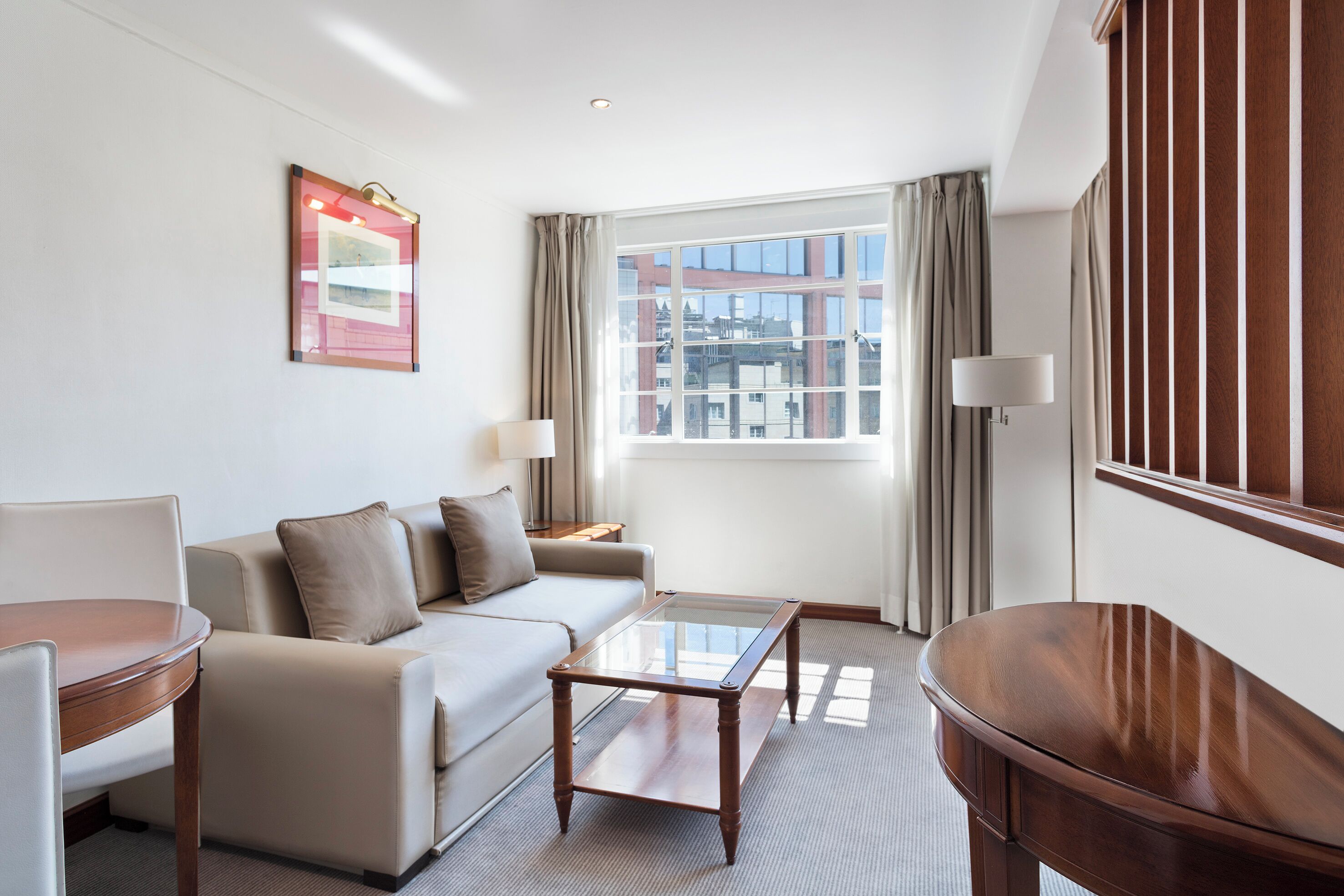 A hotel room with a sofa, a glass-top coffee table, a wooden side table, a lamp, and a window with curtains.