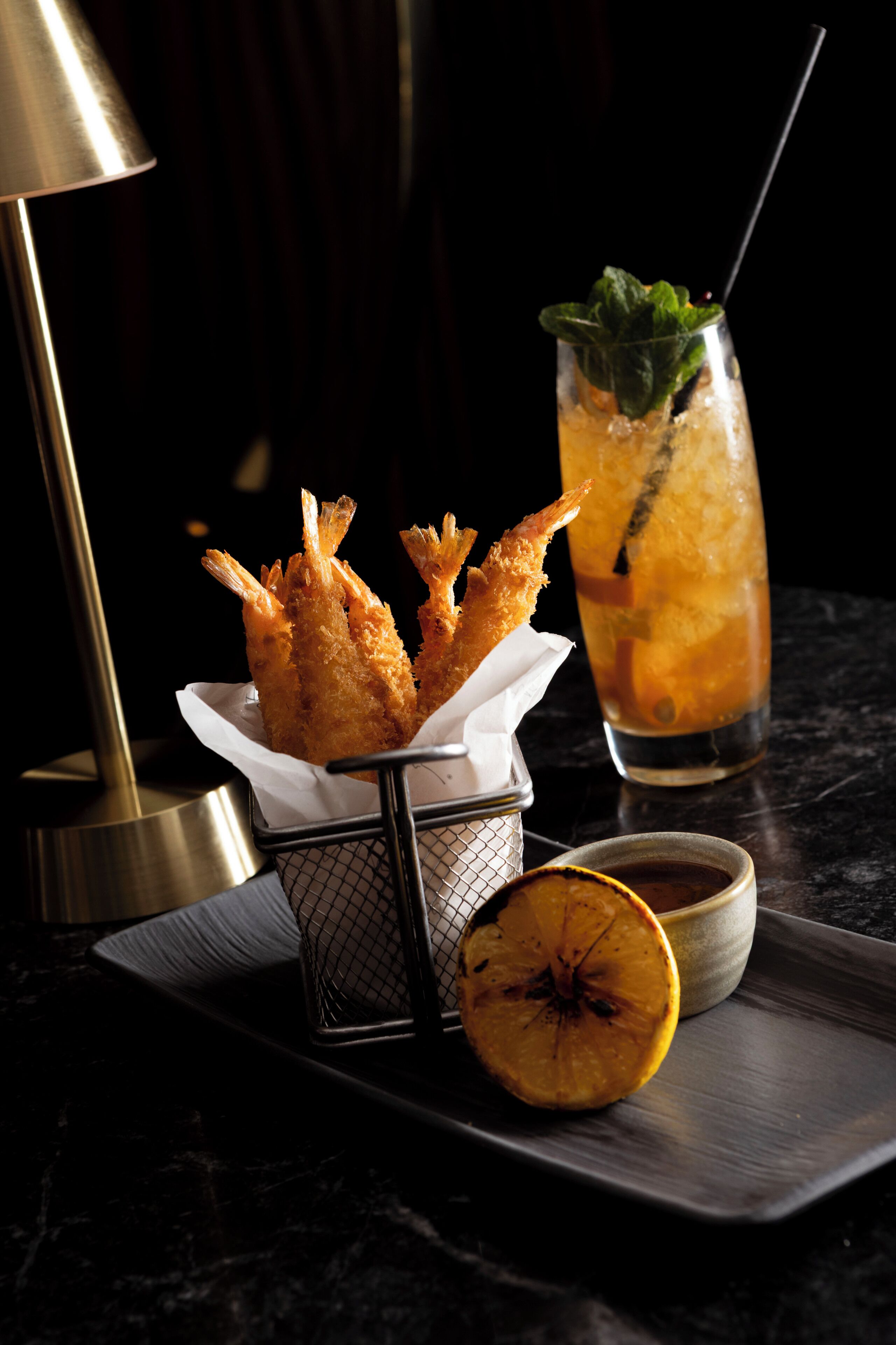 A tray with fried shrimp, a glass of iced drink with a straw, and a slice of orange on a dark surface.