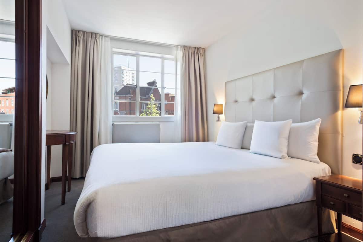 Une chambre d’hôtel avec un grand lit, un bureau, une chaise, une fenêtre avec des rideaux et une tête de lit dotée d’un luminaire.