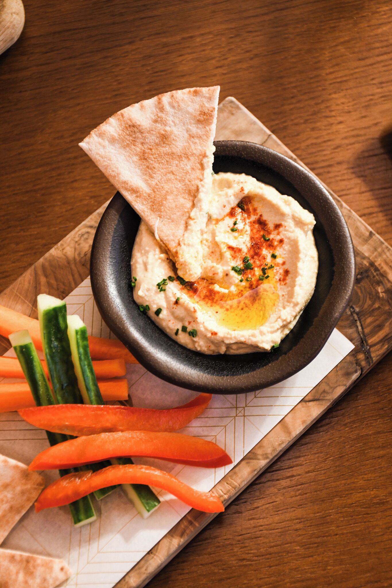 A bowl of hummus with pita bread and vegetables on a wooden board.