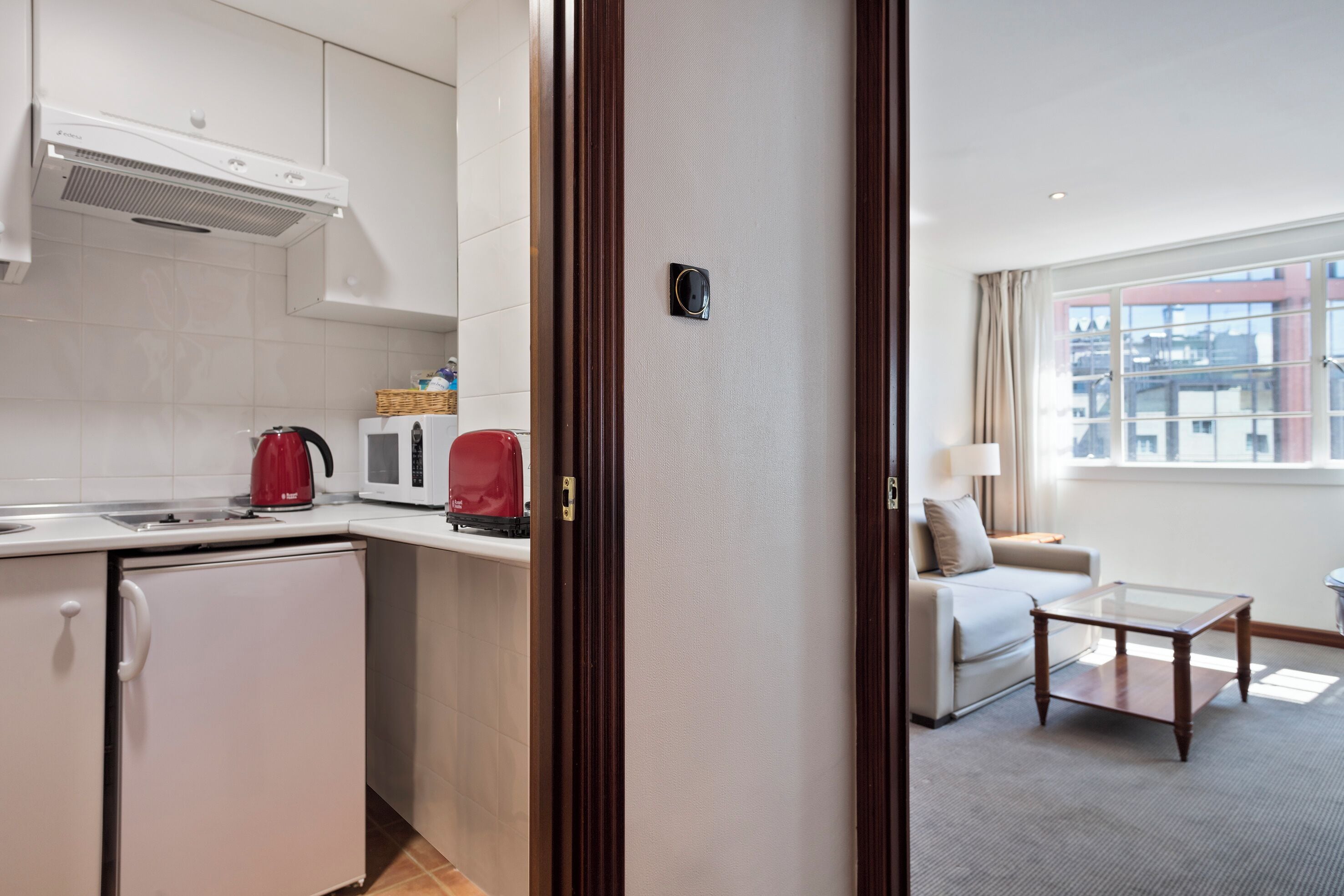 A compact hotel room with a kitchenette, a glass coffee table, a sofa, and a large window.