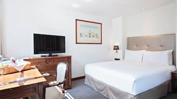 Una habitación de hotel con una cama, un escritorio con televisor, una silla, un cuadro en la pared y una mesita de noche con lámpara.
