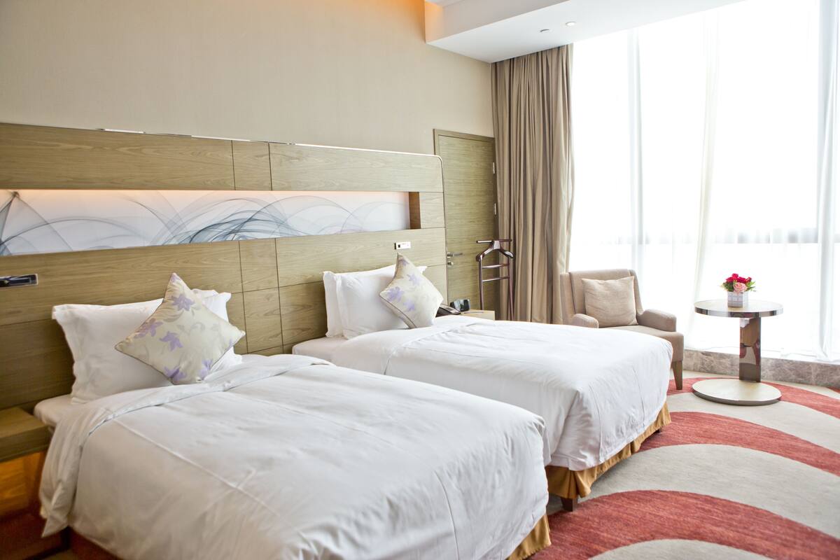 A hotel room with two beds, a round table, a chair, and a large window with curtains.