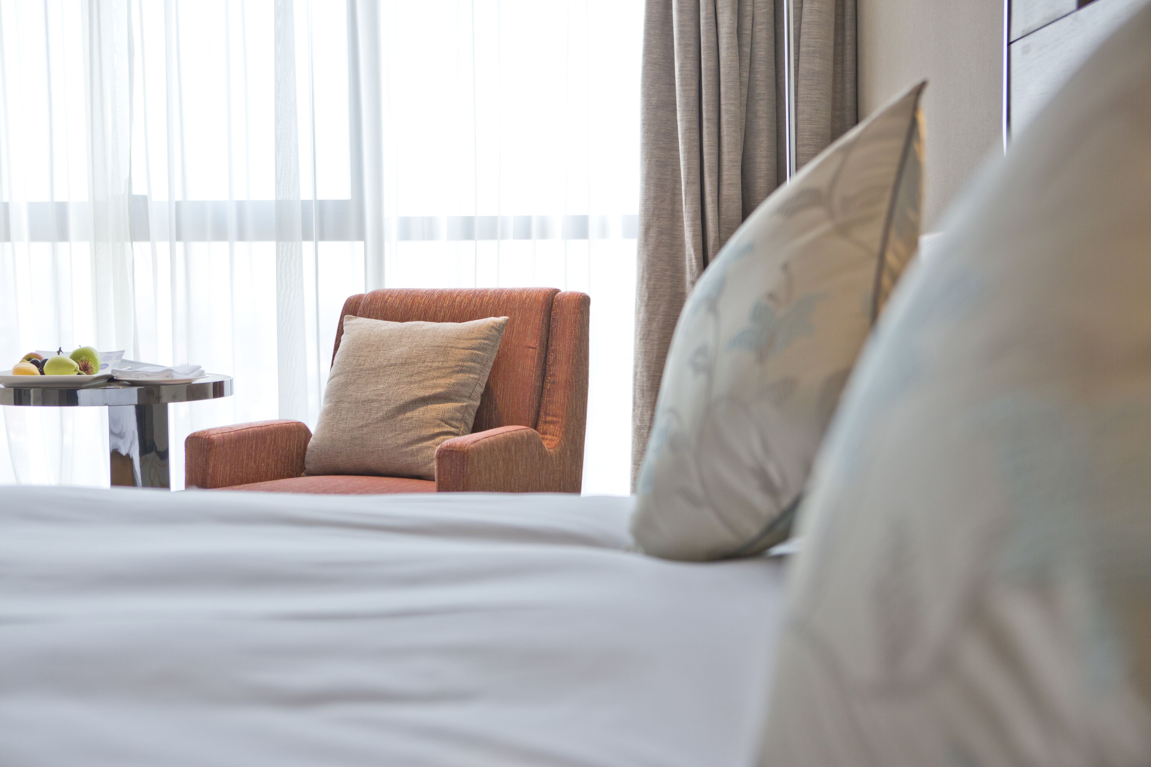 A hotel room with a bed, a chair, a small table with fruits, and a window with curtains.