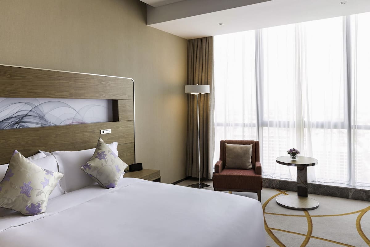 A hotel room with a large bed, a chair, a small table, and a view of the city through large windows.