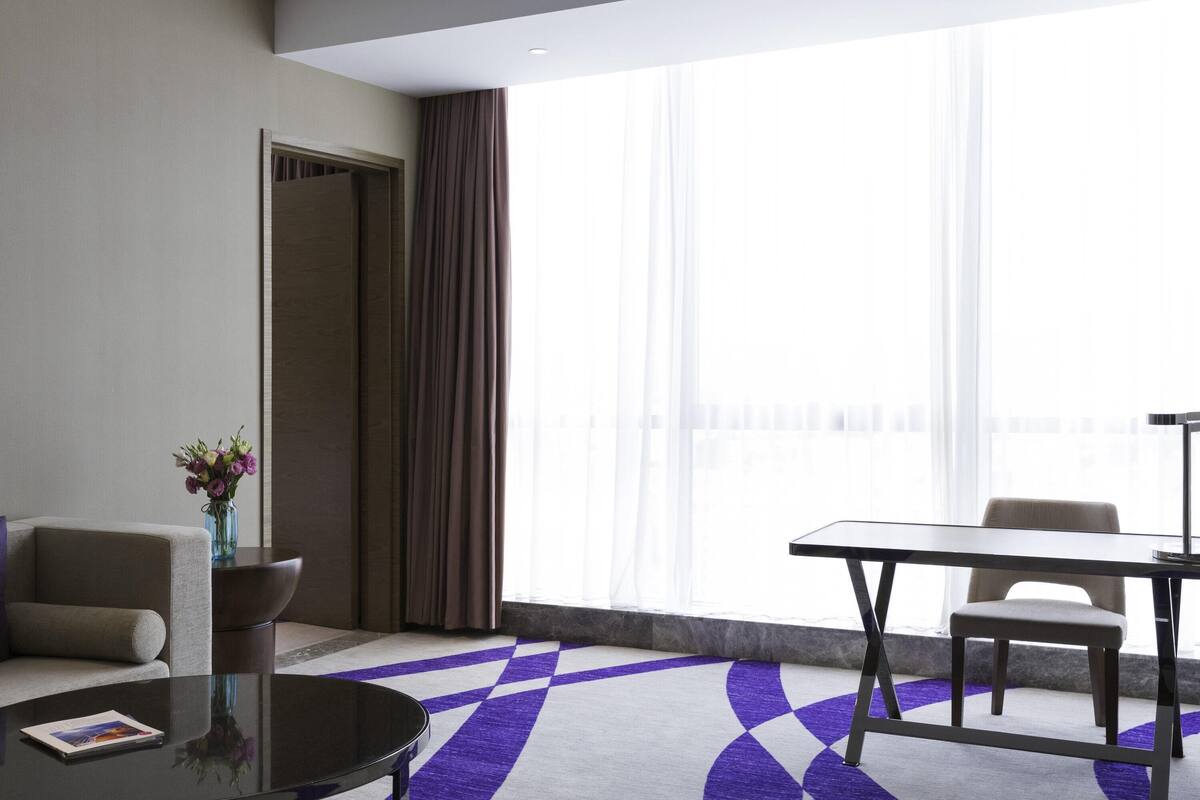 A modern hotel room with a sofa, a dining table, and a view of the city through large windows.