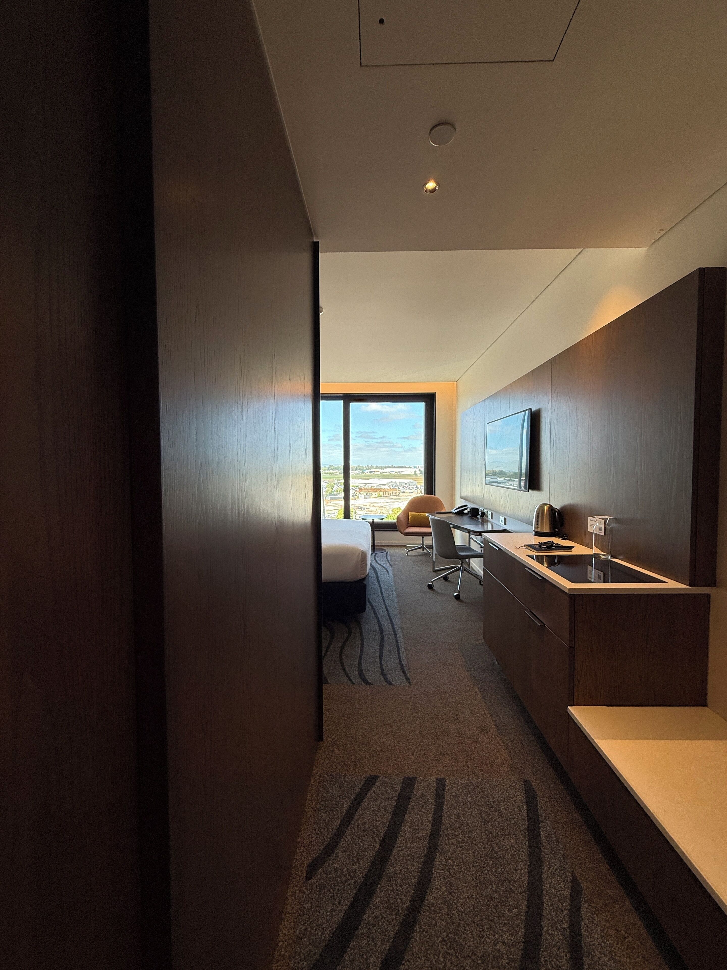 A modern hotel room with a large window, a bed, a desk, and a kitchenette.
