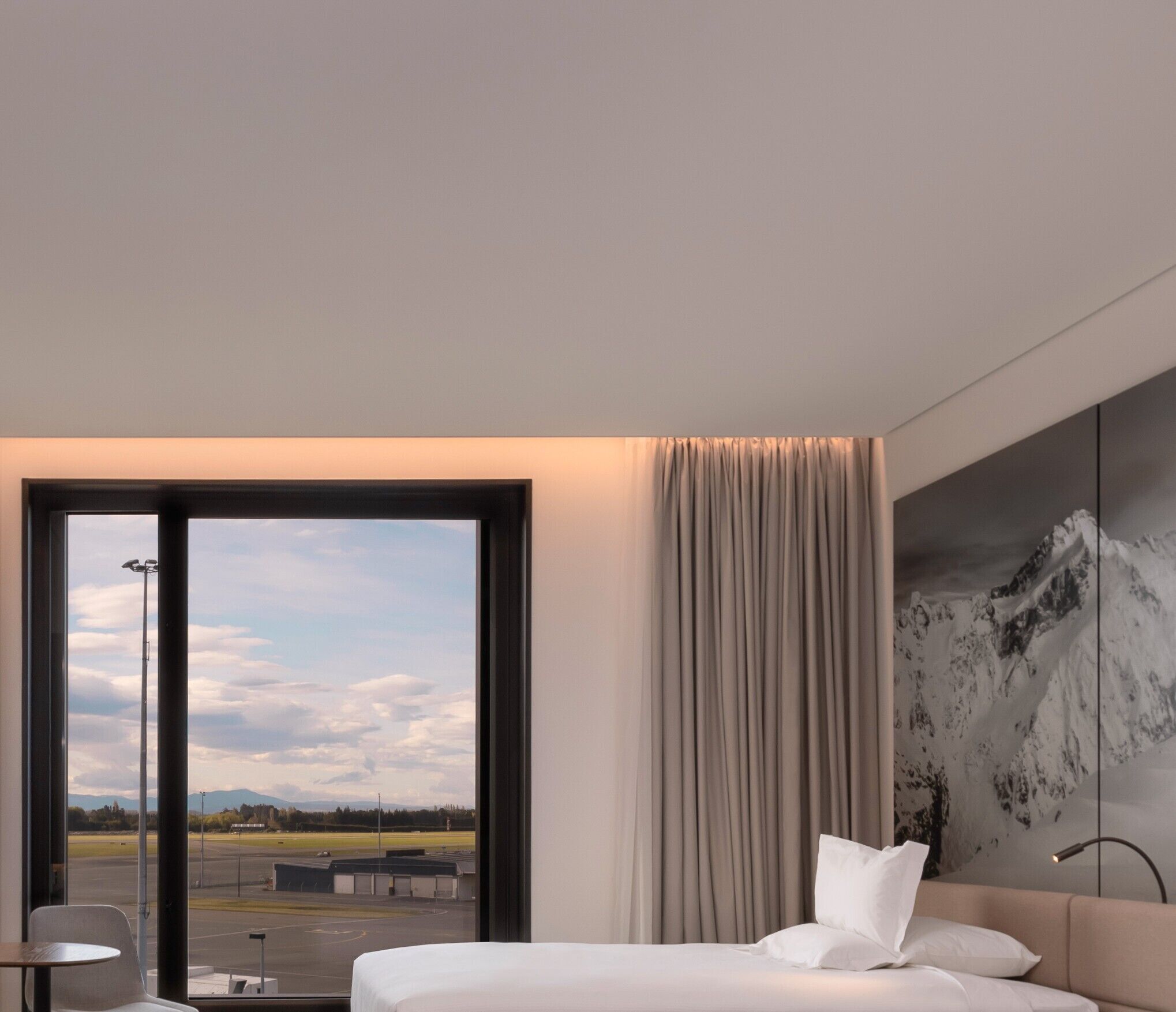 A modern hotel room with a large bed, a view of an airport, and a mountain landscape on the wall.