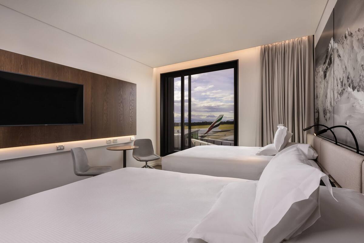 Una habitación de hotel moderna con dos camas, un televisor de pantalla plana montado en la pared, una mesita con silla y un amplio ventanal con vistas al aeropuerto.