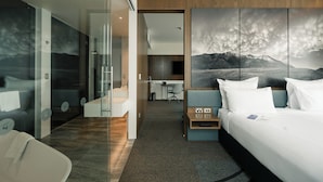 A modern hotel room with a large bed, a desk, and a glass partition.