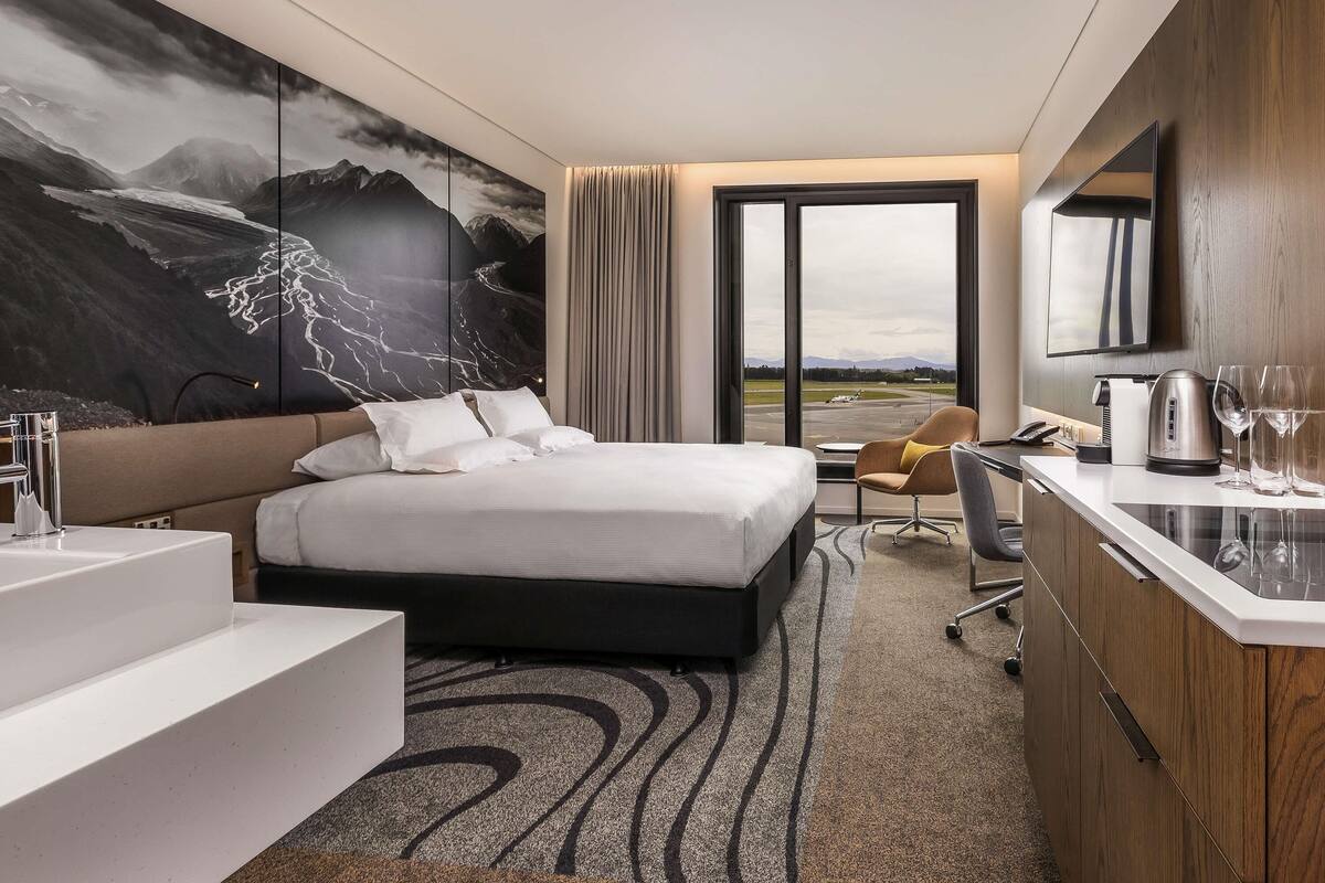 A modern hotel room with a large bed, a desk with a kettle, a chair, and a view of the airport.