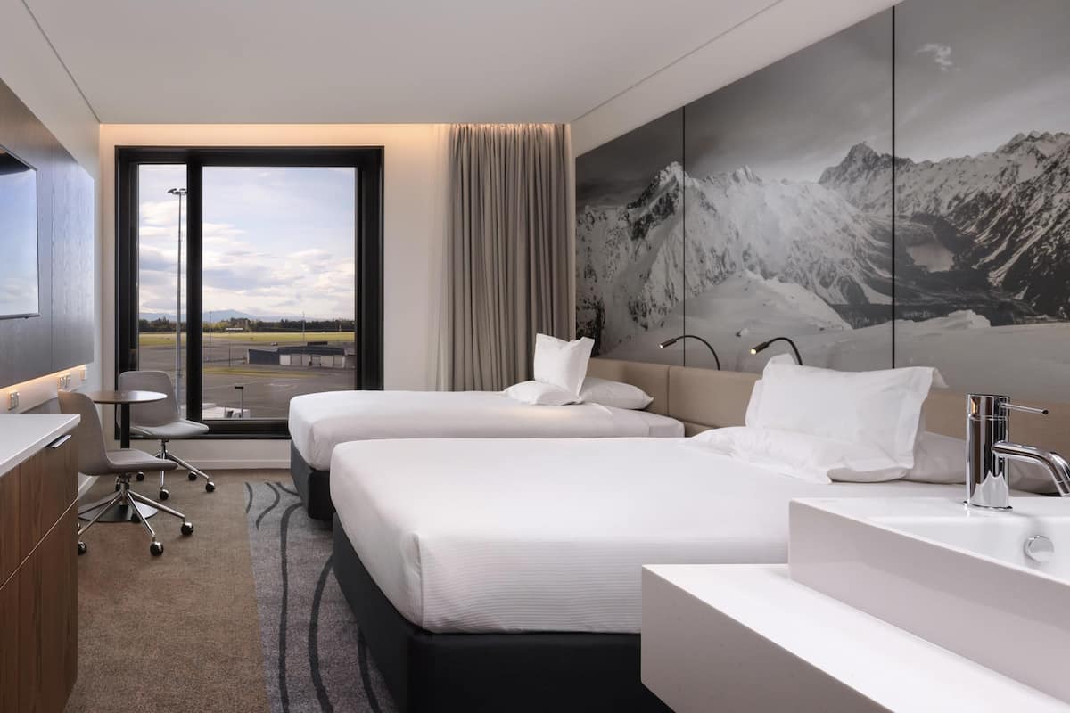 Habitación de hotel con dos camas, un escritorio, un ventanal grande con vista a un estacionamiento y un baño moderno.