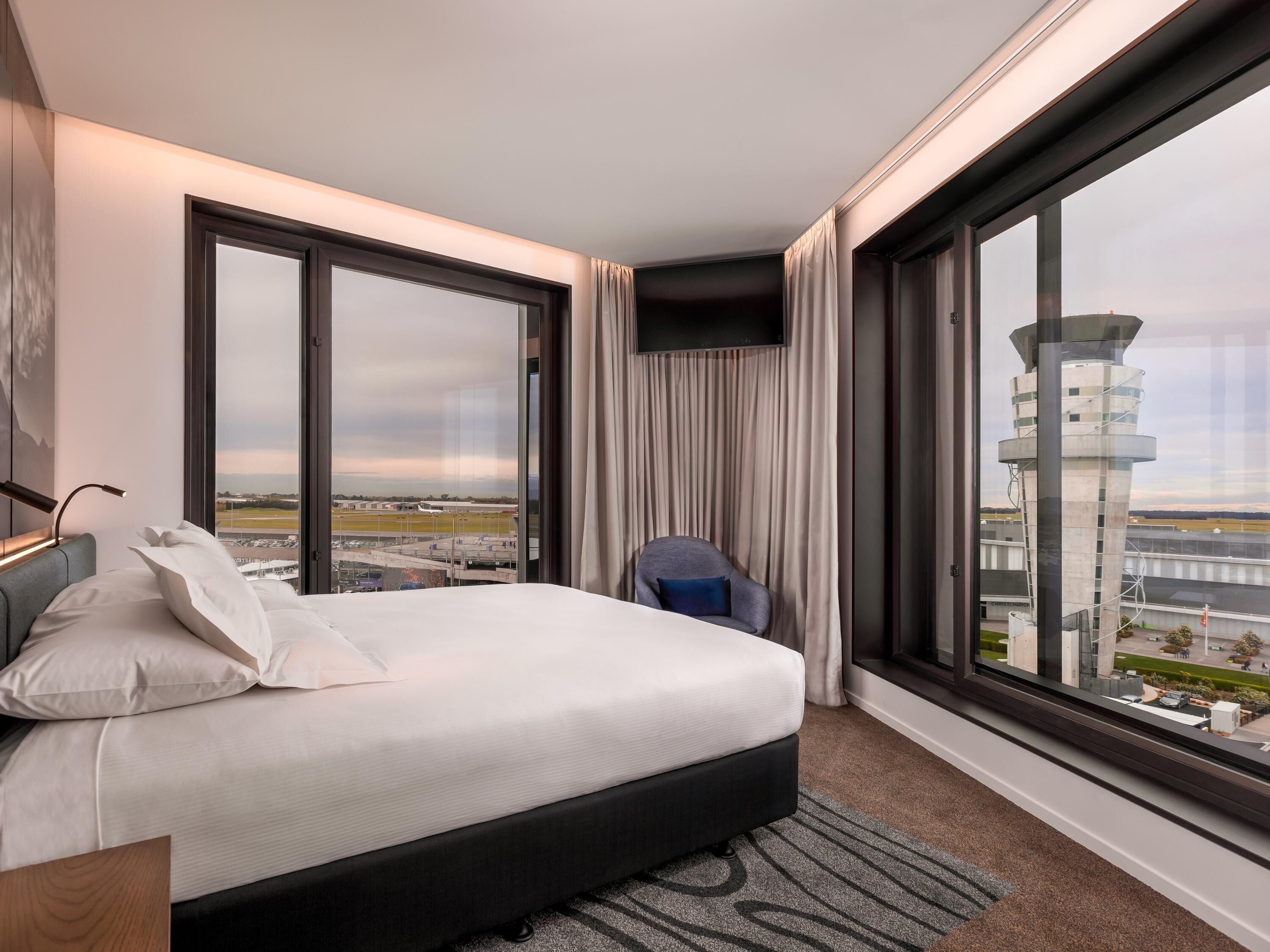 A hotel room with a large bed, a chair, a TV, and a view of an airport and tower.