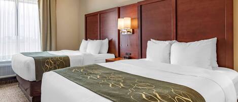 A hotel room with two beds, beige walls, a wooden headboard, a nightstand with a lamp, and a window with curtains.