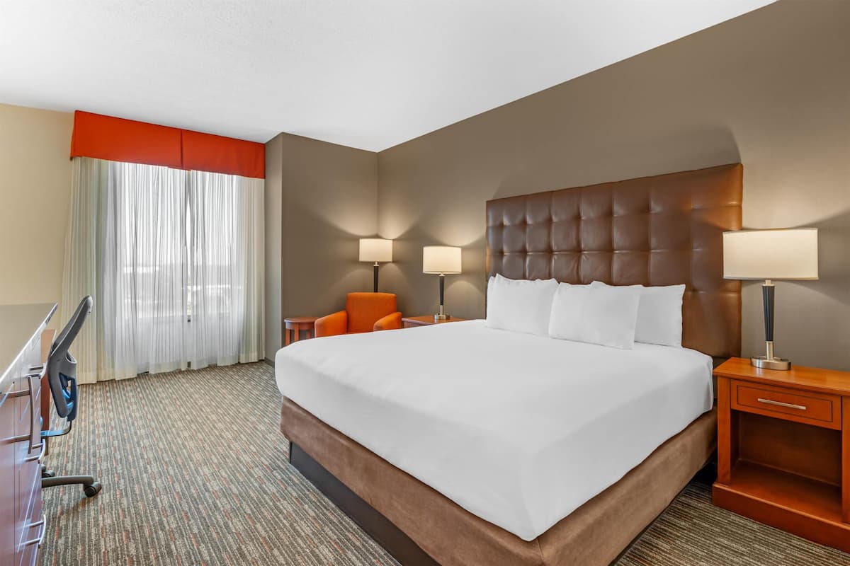 Une chambre d’hôtel avec un grand lit, deux lampes de chevet, un fauteuil orange, un bureau avec une chaise et une fenêtre avec des rideaux légers.
